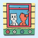 Painting on a square canvas of 2 cartoon cats, one white and one orange, sitting in the window sill of a yellow house with a green flower hedge in front of it.