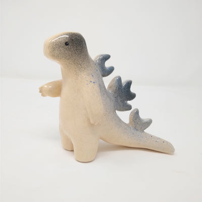 Ceramic sculpture of Godzilla, with mostly smooth body shapes and only eyes rendered on its face. It has spikes along its back and has one hand extended out slightly. Colors are mainly cream with blue and black specks centralized on its back.