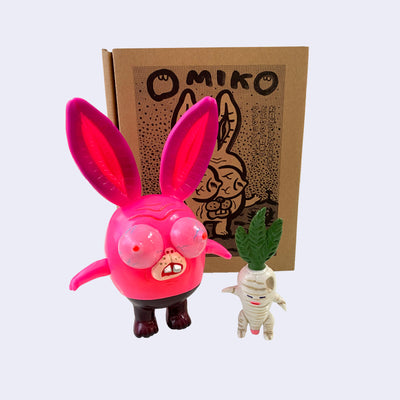 Neon pink vinyl figure of a strange looking rabbit, with female genitalia for ears, boobs for eyes and short body. It stands next to a short white daikon, with male genitalia. They stand in front of their product packaging.