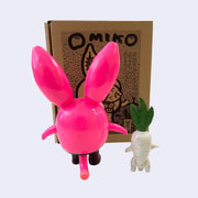 Neon pink vinyl figure of a strange looking rabbit, with female genitalia for ears, boobs for eyes and short body. It stands next to a short white daikon, with male genitalia. They stand in front of their product packaging. Back view.