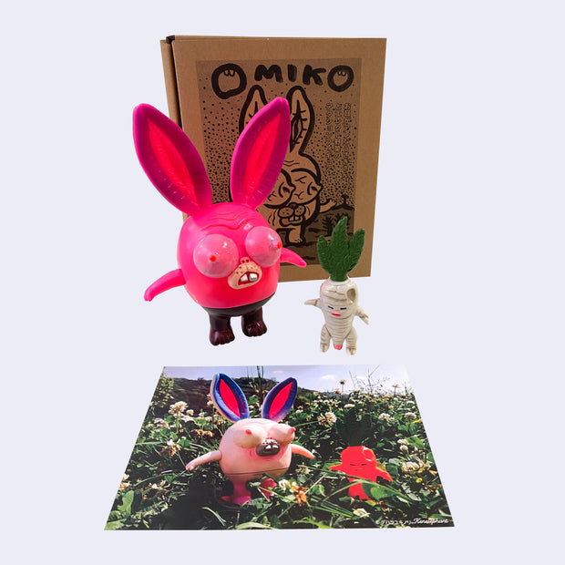 Neon pink vinyl figure of a strange looking rabbit, with female genitalia for ears, boobs for eyes and short body. It stands next to a short white daikon, with male genitalia. They stand in front of their product packaging. A photograph of the 2 figures in the grass is below.