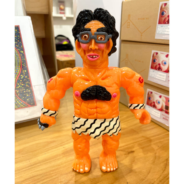 Vinyl figure of a buff, mostly nude man with black hair and glasses. He wears a striped loin cloth with matching wristbands and a set of spiked metal knuckles.