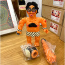 Vinyl figure of a buff, mostly nude man with black hair and glasses. He wears a striped loin cloth with matching wristbands and a set of spiked metal knuckles. At his feet is a head and a spiked club.