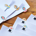 White paper envelopes with colorful illustrated clip-on tabs arranged on a wooden surface.