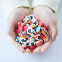 A pair of hands holding a pile of small, colorful sushi-themed erasers shaped like nigiri, salmon, and maki on a clean white background.