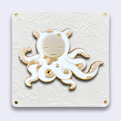 Die cut enamel pin designed by James Jean, of a white octopus outlined in gold with sparse gold swirl patterning, and a circular human like face, looking down. Its many arms are curled into itself and have light gold tentacles.