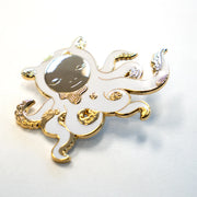 Die cut enamel pin designed by James Jean, of a white octopus outlined in gold with sparse gold swirl patterning, and a circular human like face, looking down. Its many arms are curled into itself and have light gold tentacles. Shown at an angle to display sheen.