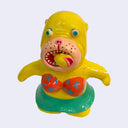 Bright yellow painted seal-like soft vinyl figure, standing with colorful eyes, an open mouth and wearing a star pattered orange bikini and green skirt. In its mouth is a fish.