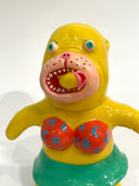 Bright yellow painted seal-like soft vinyl figure, standing with colorful eyes, an open mouth and wearing a star pattered orange bikini and green skirt. In its mouth is a fish.