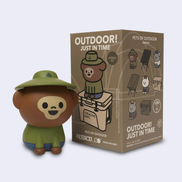 Toy figure of a bear in outdoor gear next to its packaging on a light gray background
