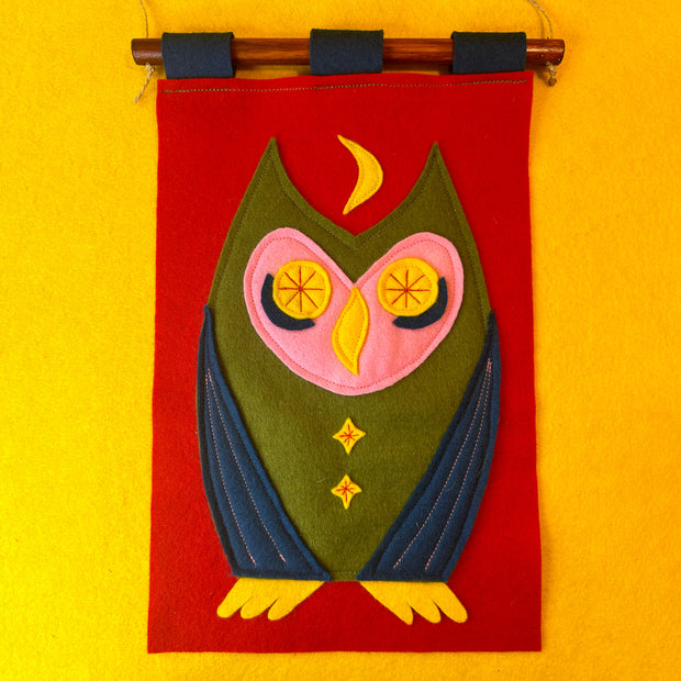 Felt wall hanging with a colorful owl design on a red background with yellow accents, hanging against a yellow wall.