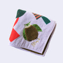 Light purple square towel with imagery of stylized green and red trees, under a small green crescent moon. A fluffy brown owl stands under the trees. Close up to show embroidered detailing.