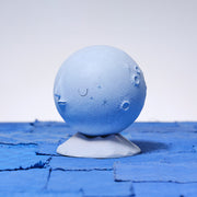 Rounded carved wooden sculpture of a light blue moon with a closed eye, calm straight faced expression. It rests atop a white, semi mountainous pedestal.
