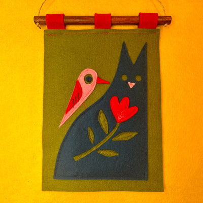 Felt wall hanging with a pink bird, navy blue cat, and red flower on a green background, hanging against a yellow wall.