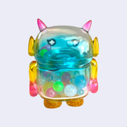 Pastel rainbow colored soft vinyl figure with colorful round beads inside. Figure is shaped like a smaller Big Boss Robot, with a bigger head than normal and two black eyes with sparkles as pupils. A red heart is on its upper right chest. Back view.