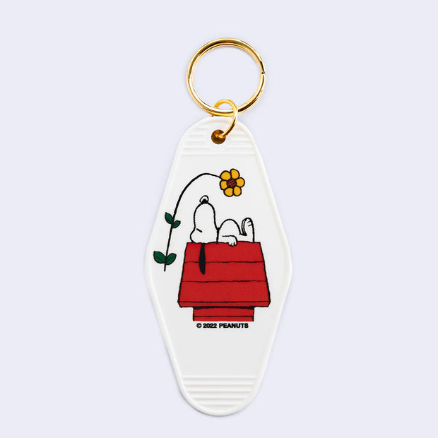 White diamond shaped key tag with rounded edges, featuring a graphic of Snoopy laying atop his iconic red dog house with a yellow flower floating over him.