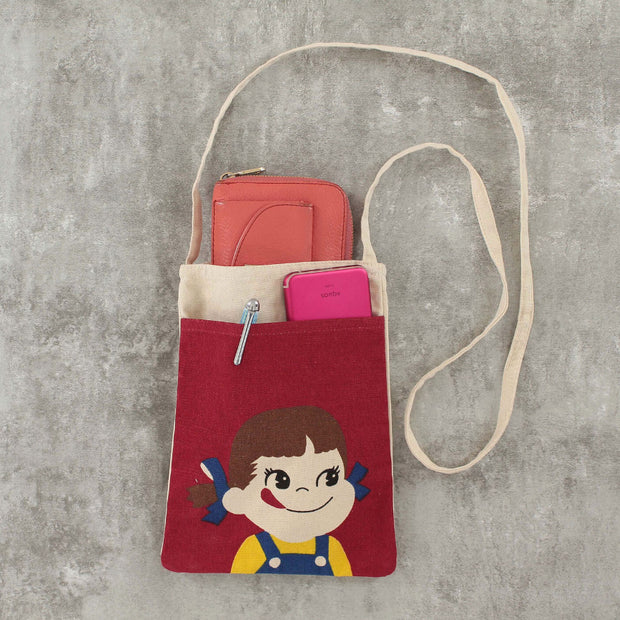 Cream colored shoulder bag with a red front pocket featuring a graphic of Peko Chan, a small cartoon girl wearing overalls and pigtails, smiling with her tongue stuck out. The bag is filled with various belongings.