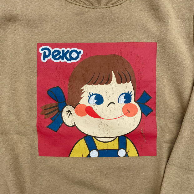 Tan colored sweatshirt with a square graphic of Peko Chan, a small smiling cartoon girl with rosy cheeks and a tongue out. She has blue bows in her hair and wears a yellow shirt and jean overalls. The graphic is weathered purposefully.