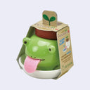 Small planter shaped like a green frog with a pink fabric tongue sticking out. It is encased in paper packaging with information on the side.