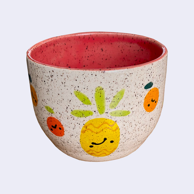 Ceramic bowl with spotted finishing and an earthy cream exterior and pinkish red interior. On the outside are painted on cartoon style citrus and a pineapple, with simple expressions.