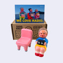 Vinyl figure of an old woman sitting with a radio on her lap. She sits next to a pink chair. Behind the figures is their product packaging and a postcard.