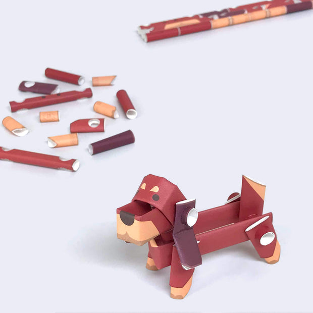 Paper sculpture of a brown dachshund, made out of folded paper pipes. It stands next to its paper pipe parts.