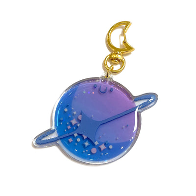 Die cut semi transparent keychain of a purple and blue ringed planet, with a small smiling face. It has sparkle designs and contains glitter within. Keychain is attached to gold moon shaped hardware.
