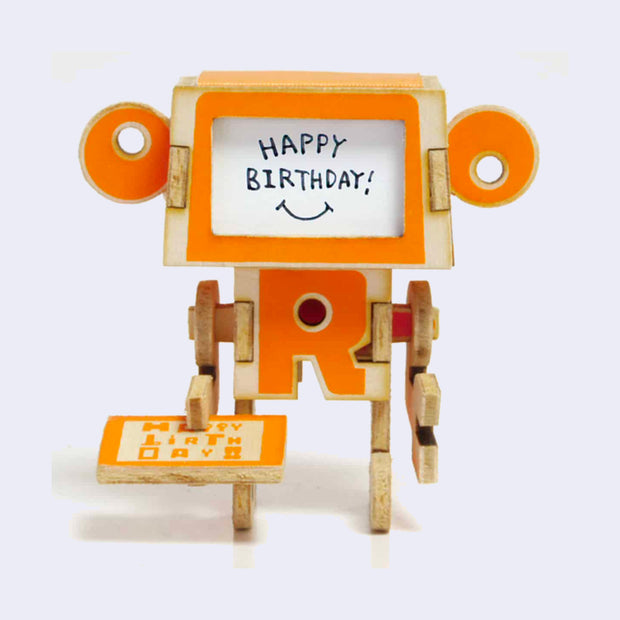Thin wooden paper sculpture of an orange robot holding a small card that reads "happy birthday." Where its face is says "happy birthday!"
