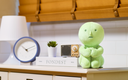 Green plush toy on a shelf with a clock, plant, and books in the background