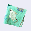 Teal green square towel with illustrations of stylized pointy green and blue mountains, with rocks. A fluffy cream colored polar bear stands at the bottom. Close up.