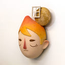 Sculpture of a person's head with a solemn yet sweet expression. They have orange hair and atop their head is a moon with a window.