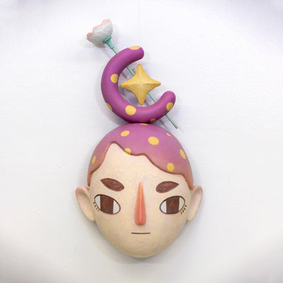 Sculpture of a person's head with a solemn yet sweet expression. They have purple and yellow polka dot hair and atop their head is a crescent with a star and a flower.