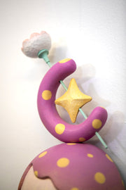 Sculpture of a person's head with a solemn yet sweet expression. They have purple and yellow polka dot hair and atop their head is a crescent with a star and a flower.