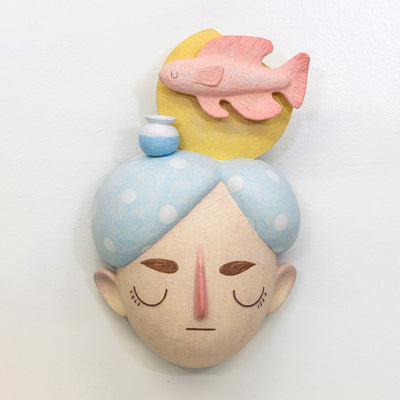 Sculpture of a person's head with a solemn yet sweet expression. They have white and blue polka dot hair and atop their head is a fish with a small vase and a yellow sun.