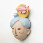 Sculpture of a person's head with a solemn yet sweet expression. They have white and blue polka dot hair and atop their head is a fish with a small vase and a yellow sun.