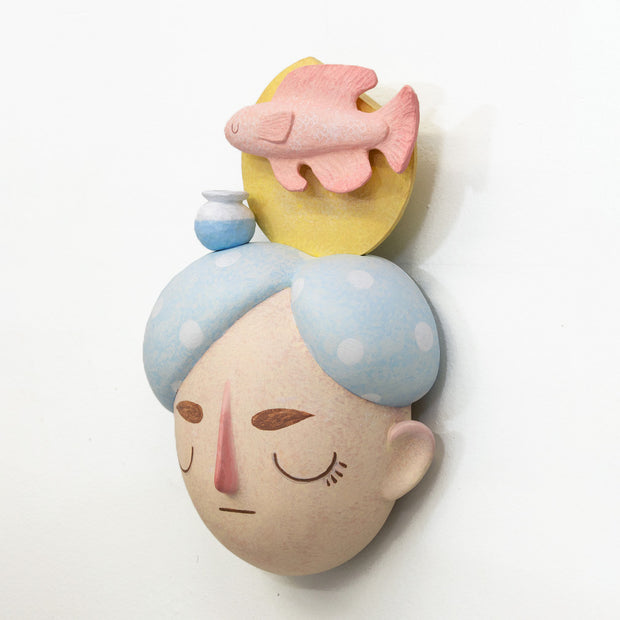 Sculpture of a person's head with a solemn yet sweet expression. They have white and blue polka dot hair and atop their head is a fish with a small vase and a yellow sun.
