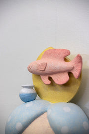 Sculpture of a person's head with a solemn yet sweet expression. They have white and blue polka dot hair and atop their head is a fish with a small vase and a yellow sun.