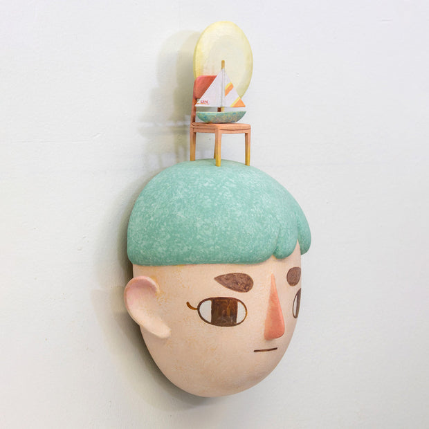 Sculpture of a person's head with a solemn yet sweet expression. They have teal hair and atop their head is a chair with a small sailboat and a large yellow moon.