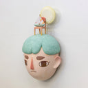 Sculpture of a person's head with a solemn yet sweet expression. They have teal hair and atop their head is a chair with a small sailboat and a large yellow moon.