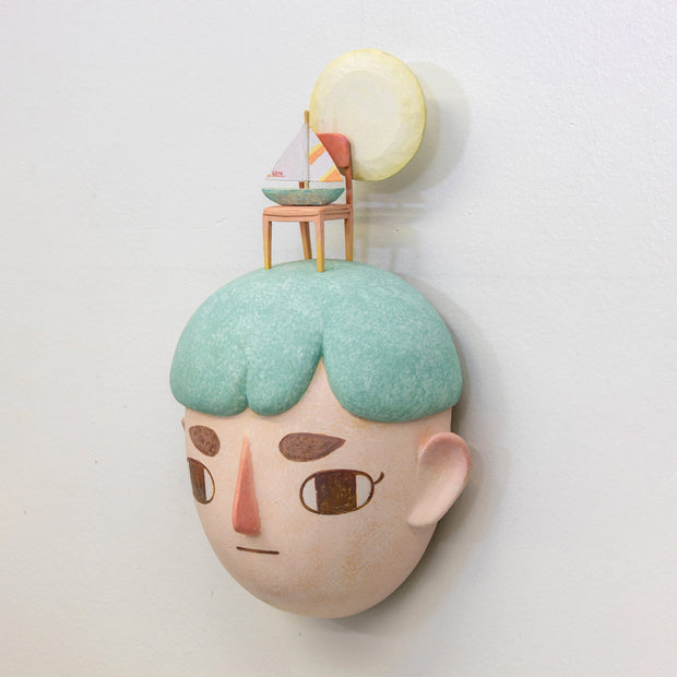 Sculpture of a person's head with a solemn yet sweet expression. They have teal hair and atop their head is a chair with a small sailboat and a large yellow moon.