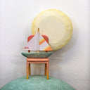 Sculpture of a person's head with a solemn yet sweet expression. They have teal hair and atop their head is a chair with a small sailboat and a large yellow moon.