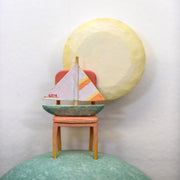 Sculpture of a person's head with a solemn yet sweet expression. They have teal hair and atop their head is a chair with a small sailboat and a large yellow moon.
