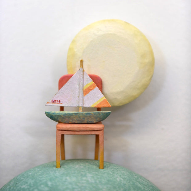 Sculpture of a person's head with a solemn yet sweet expression. They have teal hair and atop their head is a chair with a small sailboat and a large yellow moon.