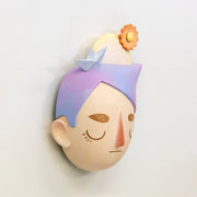Sculpture of a person's head with a solemn yet sweet expression. They have pointed purple and pink hair and atop their head is a paper sailboat, a sun and an orange flower.