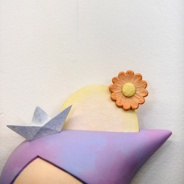 Sculpture of a person's head with a solemn yet sweet expression. They have pointed purple and pink hair and atop their head is a paper sailboat, a sun and an orange flower.