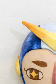 Sculpture of a person's head with a solemn yet sweet expression. They wear a helmet like a superhero with blue stripes and a large yellow arrow.