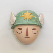 Sculpture of a person's head with a solemn yet sweet expression, They wear a military helmet with a star in front and angel wings on the side.