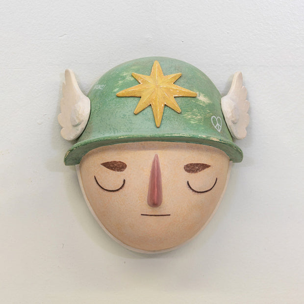 Sculpture of a person's head with a solemn yet sweet expression, They wear a military helmet with a star in front and angel wings on the side.