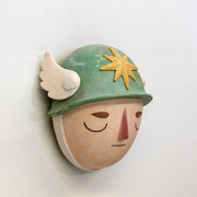 Sculpture of a person's head with a solemn yet sweet expression, They wear a military helmet with a star in front and angel wings on the side.