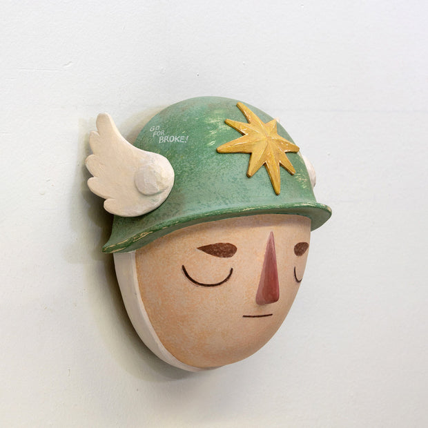 Sculpture of a person's head with a solemn yet sweet expression, They wear a military helmet with a star in front and angel wings on the side.